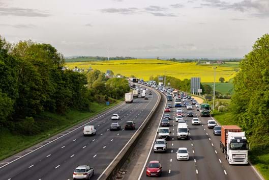 View of M1 motorway, traffic across four lanes, north and southbound in foreground, view of farm fields in background, grey sky.