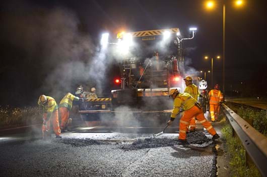 Road workers in protective clothing, resurfacing a road with asphalt and plant machinery in the background, night time.