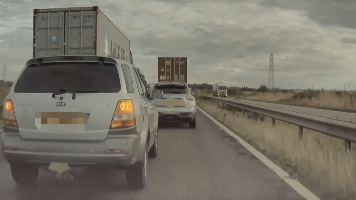 Highway scene featuring a silver SUV, with another silver SUV, and two large HGVs. The road is divided by a metal barrier, with grassy fields and power lines in the background beneath an overcast sky.