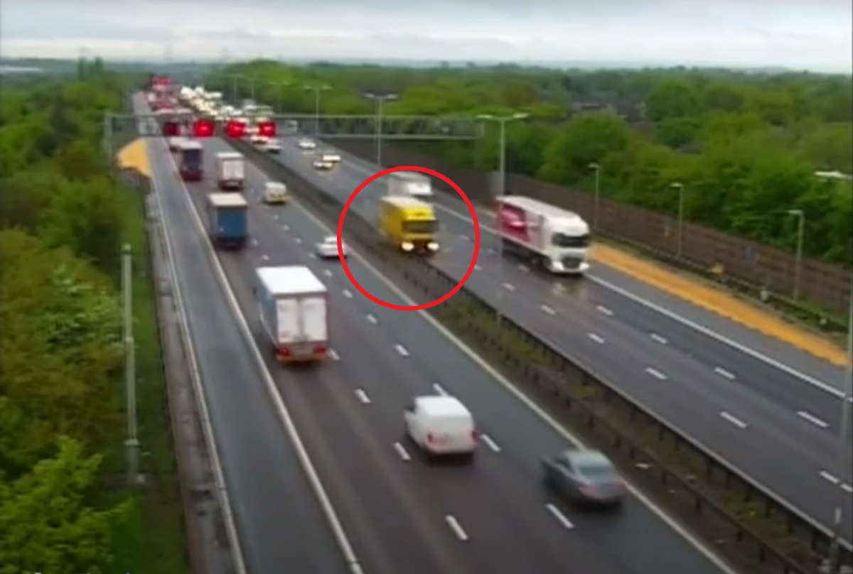 HGV travelling northbound on the carriageway to the right, veers into the third lane as it comes under the gantry before mounting the central reservation barrier and driving along the steel guard until going out of view