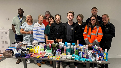 a group of office and construction workers, indoors, stood near a table which holds a collection of toiletries